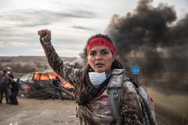 Field Medic wearing camo and a red headband that reads: "Water is Life," raising her right fist. A large wood fire behind her blocks a road. Fellow Water Protectors behind her also have fists raised.